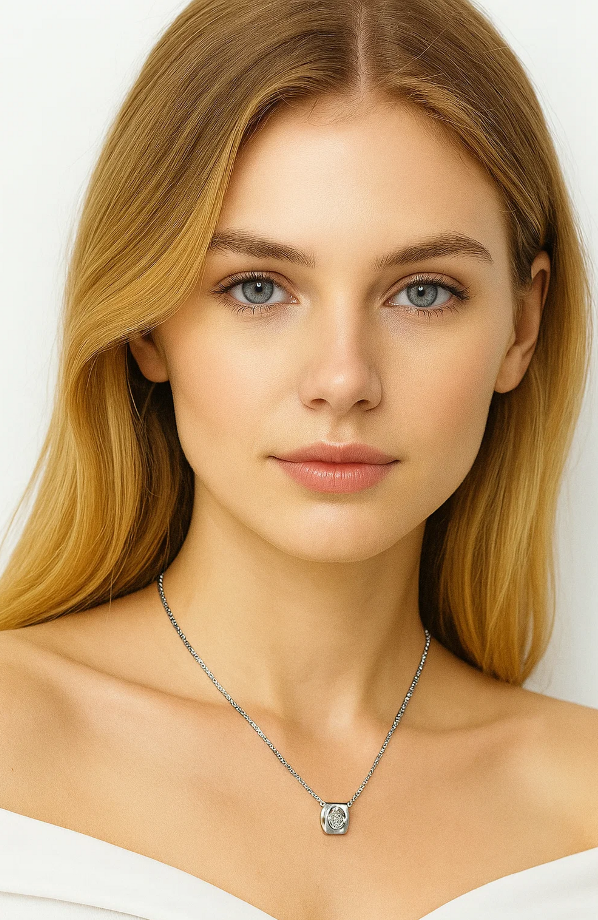 A young woman with long blonde hair and blue eyes wears the Evelyn Zirconia Heart Pendant Necklace. She has a neutral expression, is dressed in an off-shoulder white top, and stands against a plain light background.