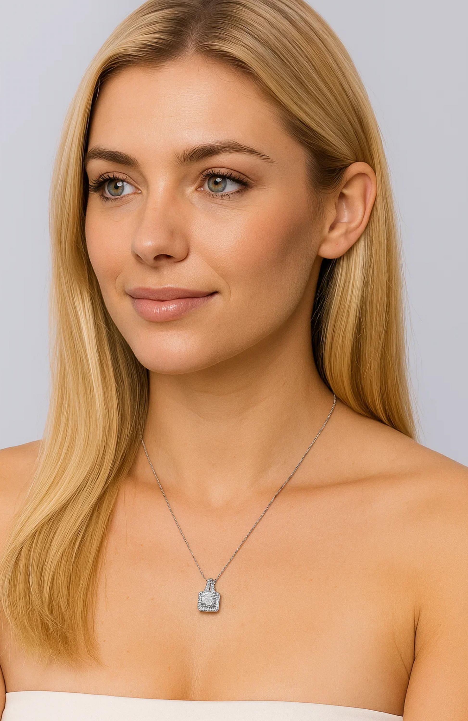 A woman with long blonde hair in a strapless top wears the Two Carat Halo Cubic Zirconia Pendant Necklace, smiling softly while gazing to the side against a light gray background.
