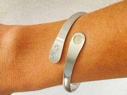 Silver cancer awareness bracelet worn on wrist, ribbon charm and round medallion
