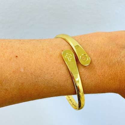 Gold bracelet cuff on wrist with cross charm and small round emblem charm