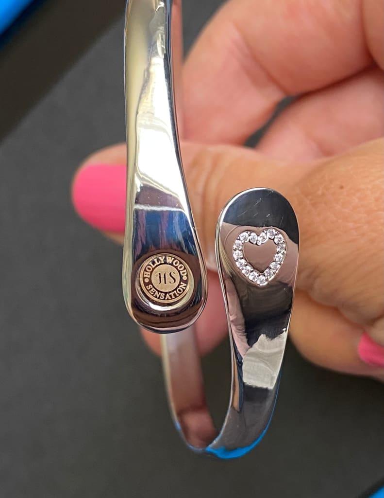 Hands holding gold and silver cuff bracelets featuring a heart charm with stones