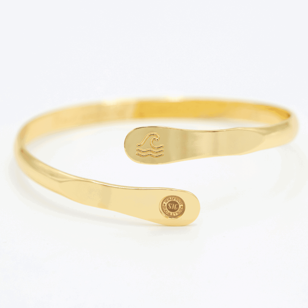 Gold wave engraved cuff bracelet angled view with wave emblem and round logo tag
