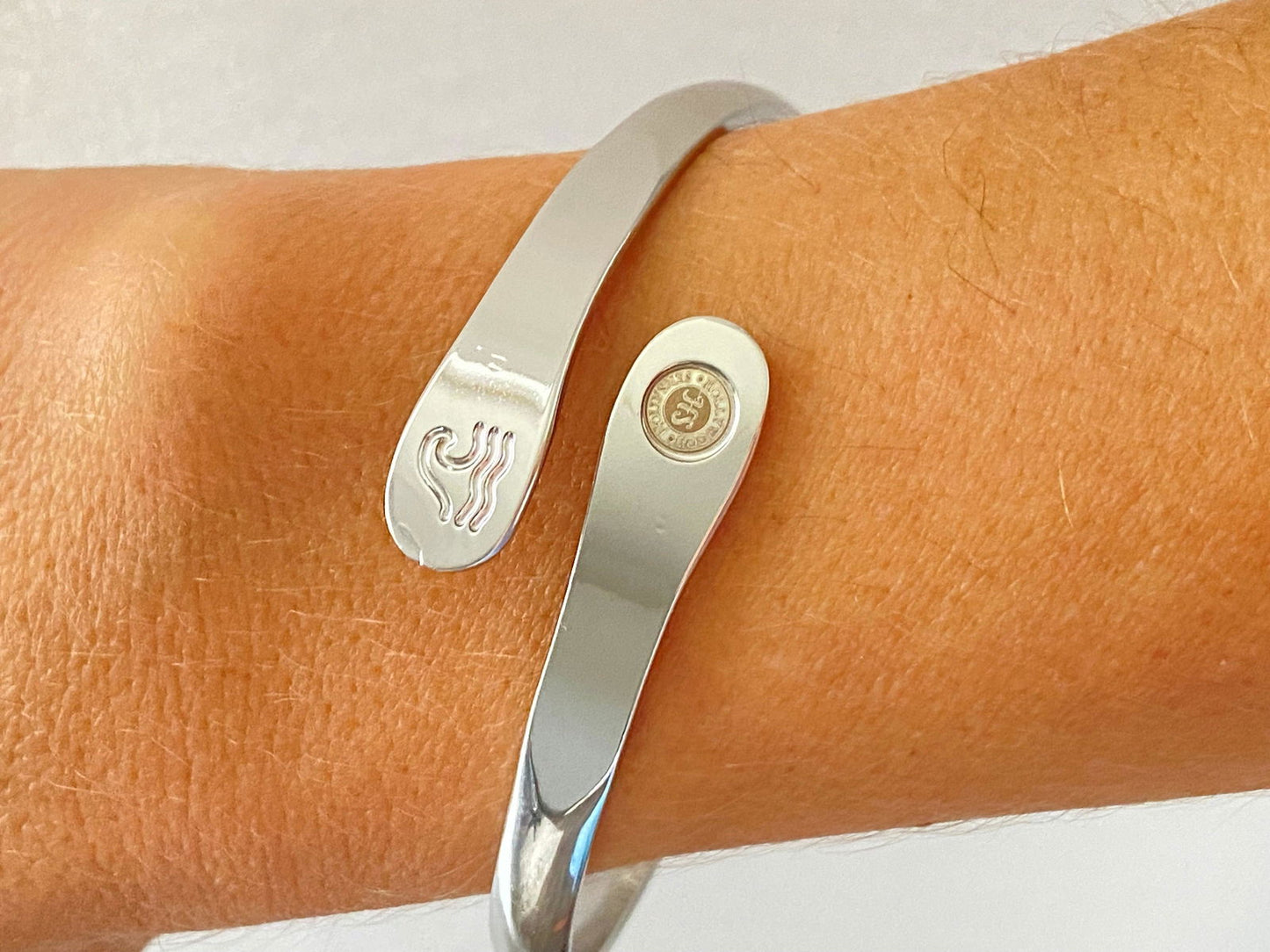 Silver wave-engraved cuff bracelet on wrist with oval wave emblem and round logo disc.