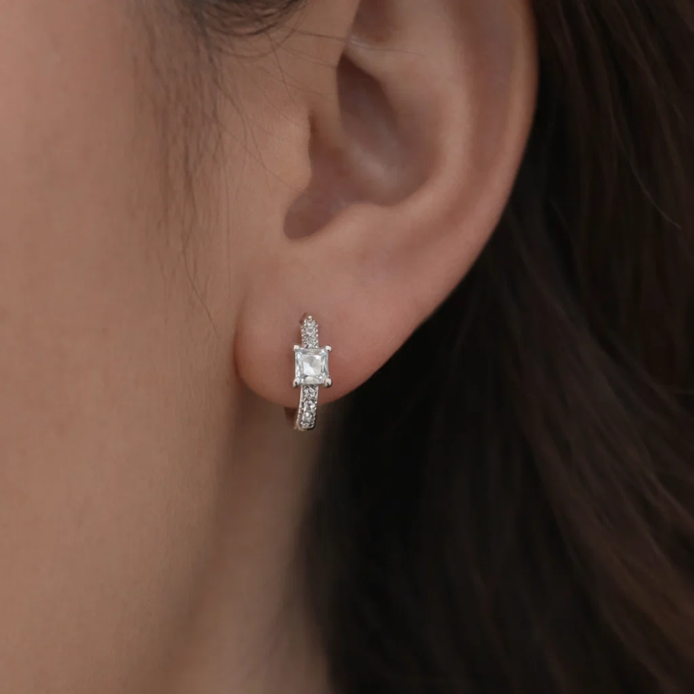 Silver square-cut huggie hoop earrings on ear with clear center stone