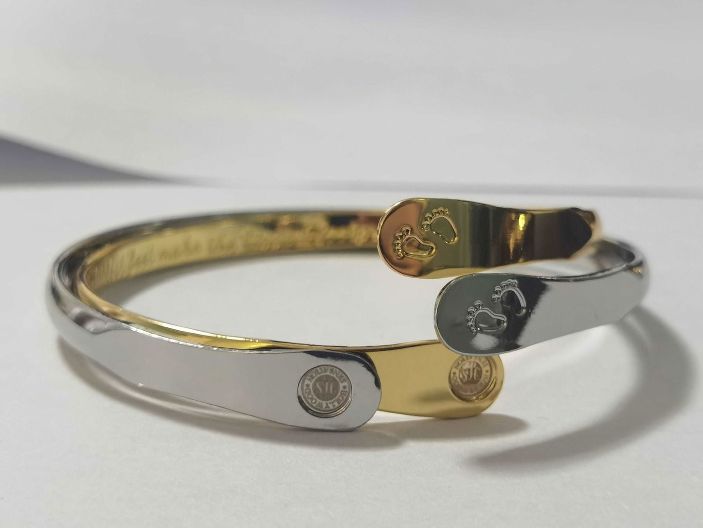 Two-tone baby footprint bangle bracelets: one silver, one gold, each with oval footprint charm.